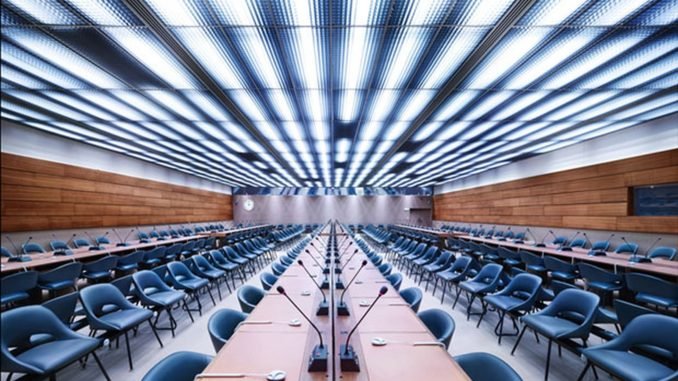 imagem de uma sala de reuniões da ONU onde acontecem os encontros do Conselho de Direitos Humanos. A sala está vazia, apenas cadeiras pretas,mesas e microfones de mesa aparecem na foto