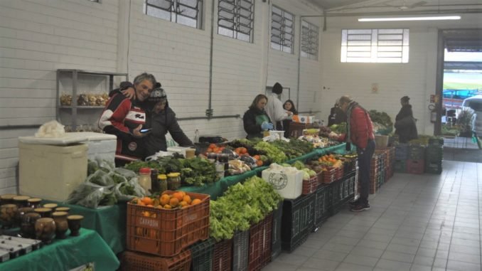 feira de alimentos da agricultura familiar com duas pessoas, um homem e uma mulher, abraçados, comercializando legumes e hostaliças